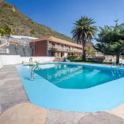 Piscina azul rodeada de palmeras y vegetación en un entorno montañoso, con una casa de estilo rústico al fondo. Ideal para relajarse y disfrutar del paisaje.