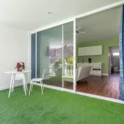 Espacio interior moderno con piso de césped sintético, mesa blanca y silla, que conecta con una sala de estar iluminada y decorada en tonos verdes. Se observa una cocina al fondo y una ventana amplia que aporta luz natural.