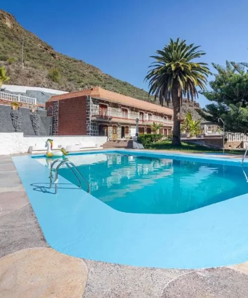Piscina azul rodeada de palmeras y vegetación en un entorno montañoso, con una casa de estilo rústico al fondo. Ideal para relajarse y disfrutar del paisaje.