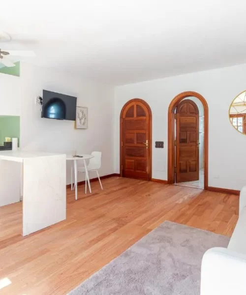Sala de estar moderna con cocina integrada, paredes verdes y muebles minimalistas. Incluye un sofá blanco, mesa de comedor, refrigerador retro y televisor en la pared. Puertas de madera y un espejo decorativo añaden calidez al espacio.