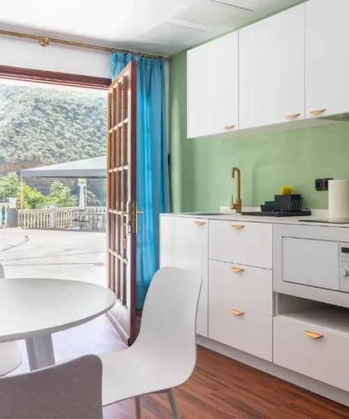Cocina moderna con paredes verdes y muebles blancos, equipada con microondas y nevera amarilla. Ventanas grandes con cortinas azules ofrecen vistas a un paisaje natural. Mesa blanca y sillas minimalistas completan el ambiente luminoso y acogedor.