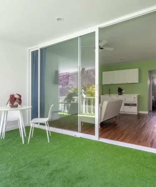 Espacio interior moderno con piso de césped sintético, mesa blanca y silla, que conecta con una sala de estar iluminada y decorada en tonos verdes. Se observa una cocina al fondo y una ventana amplia que aporta luz natural.