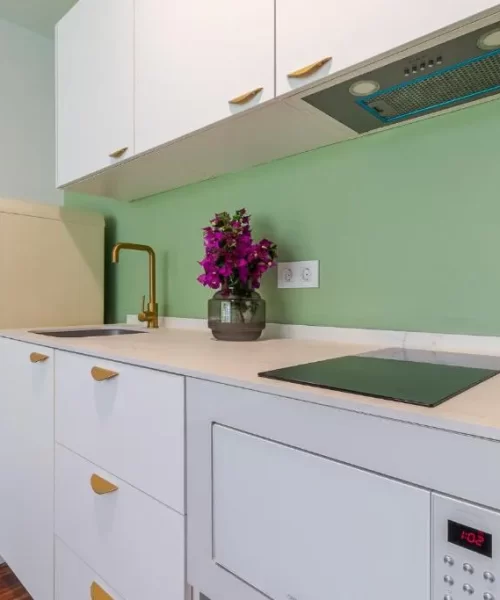 Cocina moderna con armarios blancos, fregadero, estufa y microondas, decorada con una planta de flores moradas y una nevera vintage de color crema, todo sobre un fondo verde claro.